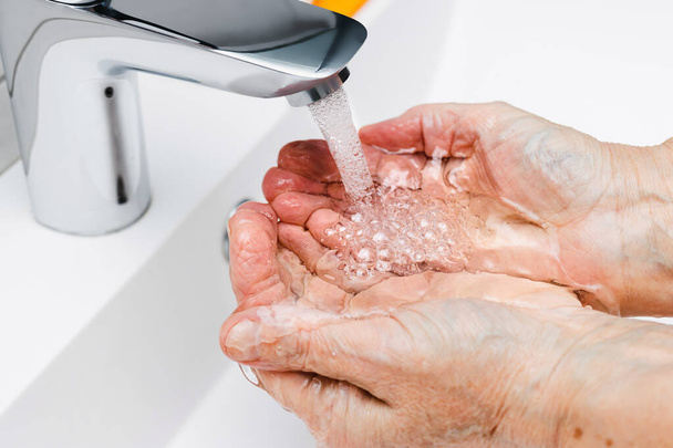 Γριά γυναίκα πλένει τα χέρια κάτω από τρεχούμενο νερό από βρύση χωρίς σαπούνι στο μπάνιο, έννοια υγιεινής. - Φωτογραφία, εικόνα