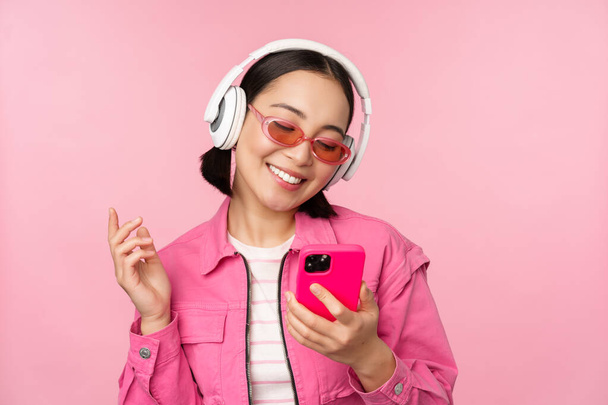 Κομψό κορίτσι από την Ασία χορεύει με smartphone, ακούει μουσική σε ακουστικά σε εφαρμογή για κινητά τηλέφωνα, χαμογελάει και γελάει, ποζάρει σε ροζ φόντο - Φωτογραφία, εικόνα