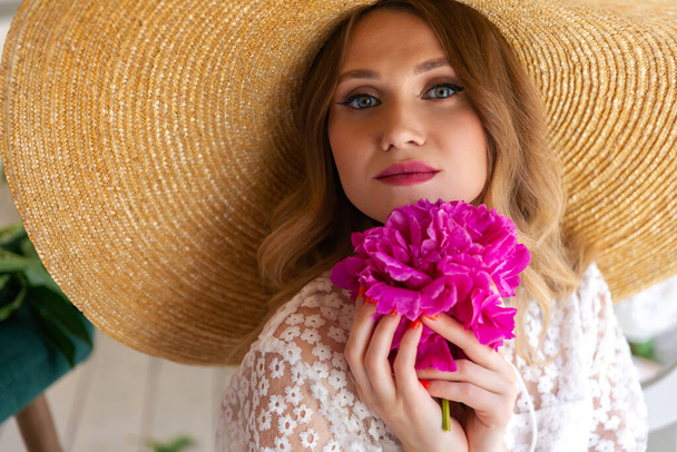 Close-up portrait of a beautiful woman in a straw hat, looking at the camera. Woman with makeup and red lips, red peony in her hands. Blonde with curled hair. Place for your text. - 写真・画像