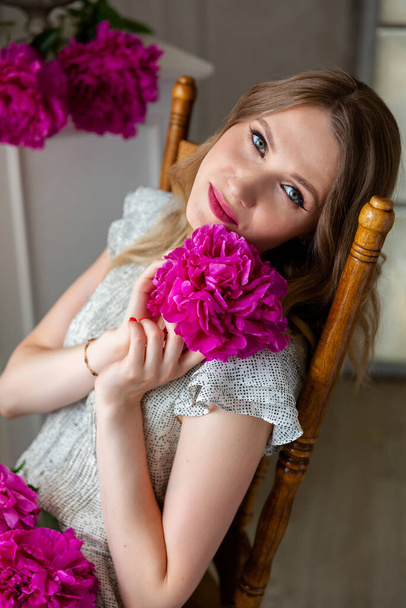 woman in white dress sitting on vintage chair, holding peony bud in her hands, looking at camera. Portrait of beautiful blonde woman. Surprise bouquet of flowers for date, mothers day, valentines day - Фото, изображение