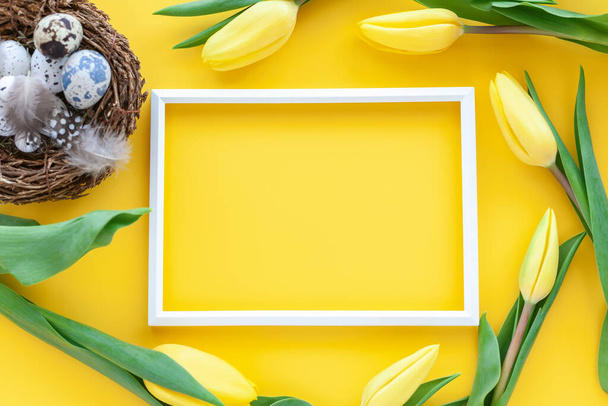 Easter holiday background with quail nest with eggs, photo frame and tulip flowers on a yellow background. Flat lay greeting card with space for text. Easter decoration, minimalist design, modern design template.  - Foto, Bild