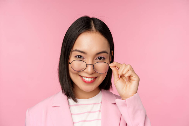 Portré ázsiai üzletasszony szemüvegben, kíváncsi a kamera és a mosolygós, professzionális eladó bámult érdeklődéssel, rózsaszín háttér - Fotó, kép