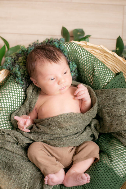 Cute little baby in a wicker basket decorated with green branches in a green knitted blanket at home on a light wooden background  - Photo, Image