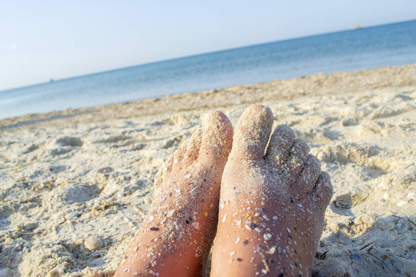 Δύο γυμνά πόδια καυκάσιας γυναίκας λερωμένης στην άμμο με κοχύλι στην αμμώδη θάλασσα - Φωτογραφία, εικόνα