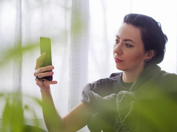 Γυναίκα με ένα τηλέφωνο στα χέρια της στο σπίτι ή στο γραφείο. Σε πρώτο πλάνο είναι μερικά φυτά σπίτι εκτός εστίασης, θολές εικόνες - Φωτογραφία, εικόνα