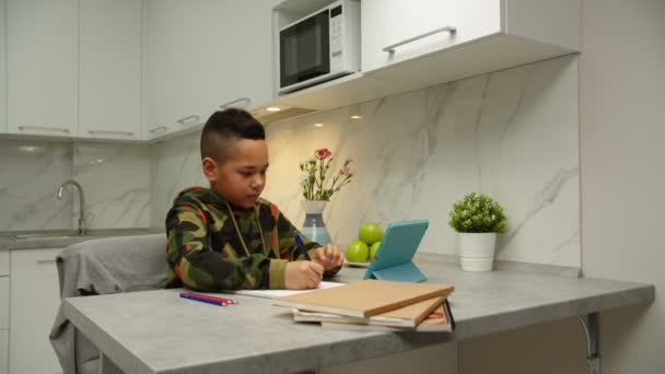 Симпатичный мальчик, пишущий в тетрадке, используя цифровой планшет во время домашнего обучения - Кадры, видео