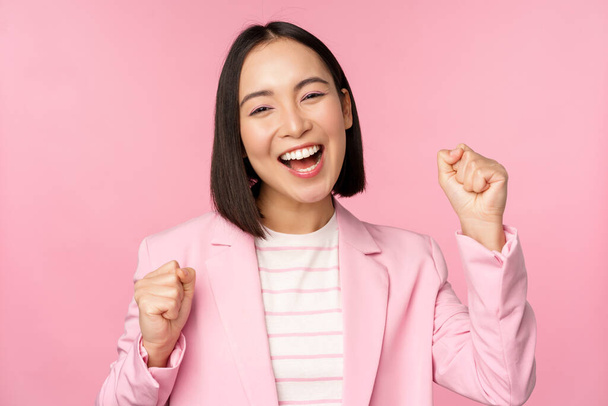Enthusiastische Verkäuferin, asiatische Unternehmensfrau sagt Ja, erreicht das Ziel und feiert, triumphiert, schaut mit Freude und lächelt, steht über rosa Hintergrund - Foto, Bild