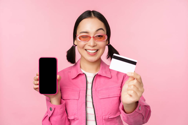 Obraz uśmiechniętej koreanki pokazującej kartę kredytową i ekran telefonu komórkowego, interfejs aplikacji smartfona, płatności online, zakupy bezstykowe, stojące nad różowym tłem - Zdjęcie, obraz