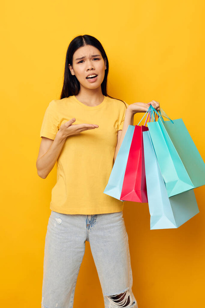 Charming young Asian woman in a yellow T-shirt with multicolored shopping bags yellow background unaltered - Фото, изображение