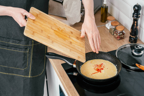 Unrecognizable man in black apron put chopped red bell pepper in omelette from wooden cutting board in frying pan. - Foto, imagen