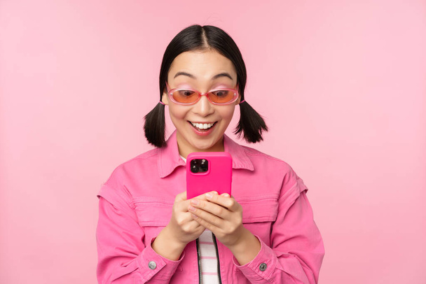 Retrato de chica asiática en gafas de sol usando smartphone. Mujer mirando el teléfono móvil, la navegación en la aplicación, de pie sobre el fondo rosa - Foto, imagen