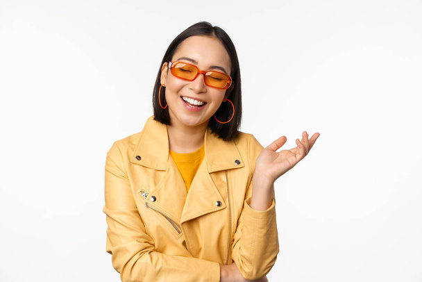 Stylowa, nowoczesna koreańska dziewczyna w okularach przeciwsłonecznych i żółtym płaszczu, śmiejąca się i uśmiechnięta, szczęśliwa, normalna ekspresja twarzy, pozująca na białym tle studia - Zdjęcie, obraz