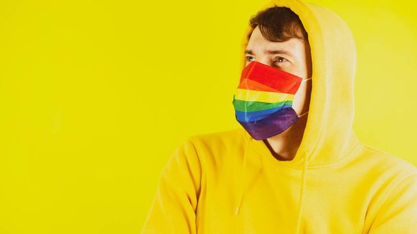 Man in protective rainbow mask on yellow background. Portrait of young male in yellow hoodie and in mask lgbt - Valokuva, kuva
