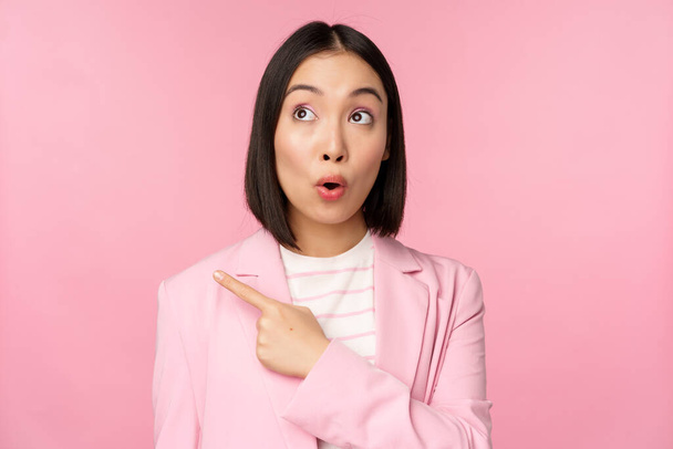 Portrait of saleswoman, korean businesswoman pointing and looking left with surprised, intrigued face expression, posing in suit over pink background - Foto, Imagem