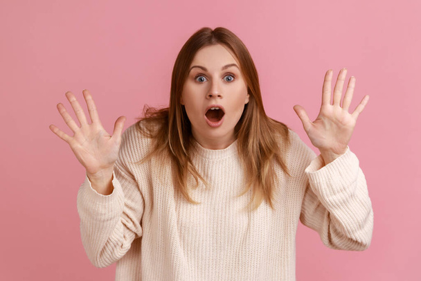 Portrait of shocked young adult blond woman standing with raised arms, big eyes and open mouth, being surprised, wearing white sweater. Indoor studio shot isolated on pink background. - Fotoğraf, Görsel