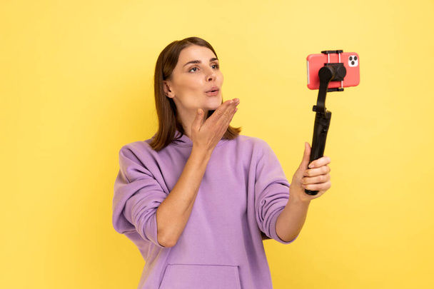 Portrait of romantic woman using cell phone and steadicam for broadcasting livestream, sending air kisses to followers, wearing purple hoodie. Indoor studio shot isolated on yellow background. - Фото, изображение