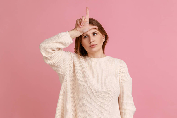 Portrait of upset blond woman standing with hand on forehead showing loser gesture, unemployed or fired from job, wearing white sweater. Indoor studio shot isolated on pink background. - Фото, изображение