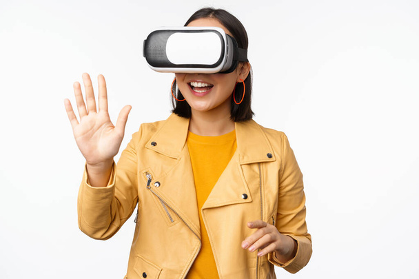 Heureuse fille asiatique dans le casque VR, disant bonjour dans le chat de réalité virtuelle, en utilisant des lunettes spéciales, debout sur fond blanc, agitant la main, disant bonjour - Photo, image