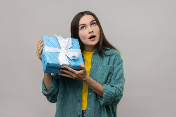 Portrait of curious woman shaking wrapped present on birthday, trying to guess what it is, looks doubtfully up, wearing casual style jacket. Indoor studio shot isolated on gray background. - Photo, image