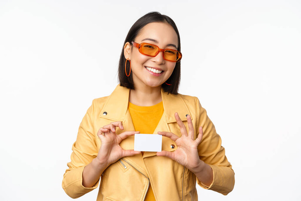 Стильна приваблива азіатська дівчина в сонцезахисних окулярах, показує кредитну картку і посміхається, стоїть щасливим на білому фоні студії
 - Фото, зображення