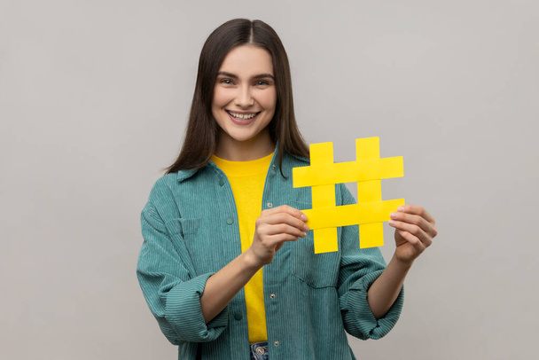 Portrait of happy beautiful woman holding hashtag symbol, promoting viral topic in social network, tagging blog trends, wearing casual style jacket. Indoor studio shot isolated on gray background. - Valokuva, kuva