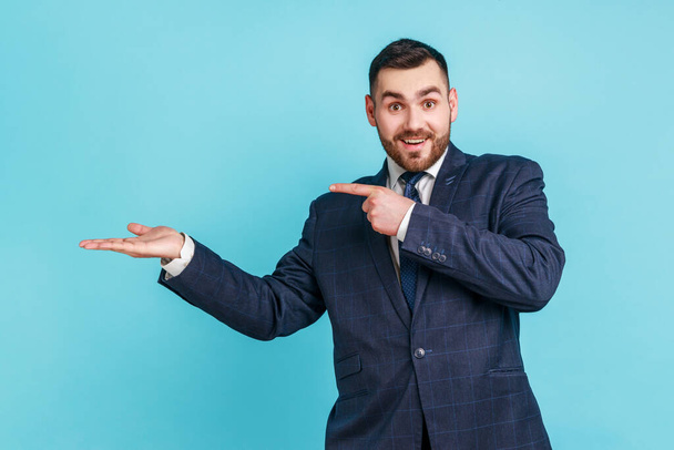 Wow, look at commercial proposal! Bearded man in suit pointing at copy space on palm, showing empty place for idea presentation, product advertising. Indoor studio shot isolated on blue background. - Φωτογραφία, εικόνα