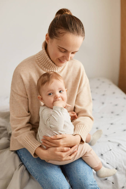 Indoor shot of attractive woman with ponytail wearing beige sweater sitting on bed with toddler daughter, mother looking at baby with love and gentle, spending time together. - Фото, зображення