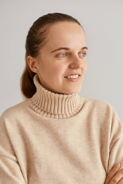 Closeup portrait of attractive dark haired woman with ponytail wearing beige sweater looking away with happy positive facial expression, female with perfect skin posing indoor. - Фото, изображение