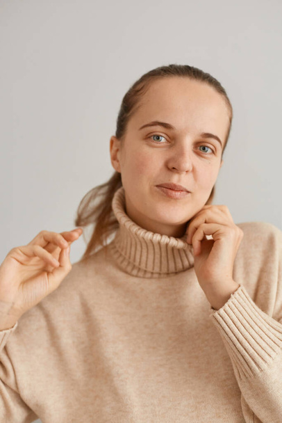 Closeup portrait of beautiful woman with ponytail wearing beige jumper looking at camera with happy positive facial expression, female keeping hands on her collar and hair. - Фото, зображення
