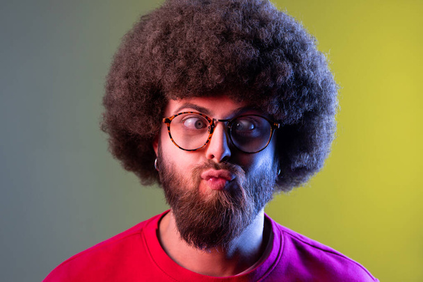 Portrait of crazy hipster man with Afro hairstyle standing with crossed eyes and looking with funny comedian face, wearing red sweatshirt. Indoor studio shot isolated on colorful neon light background - Фото, изображение