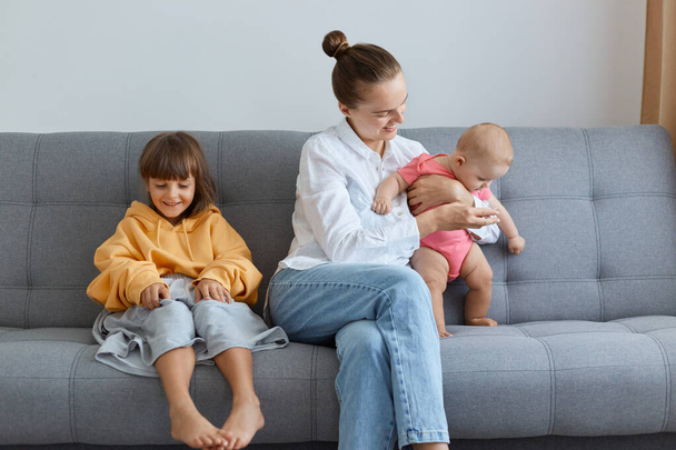 Portrait of young adult mother with her kids sitting on sofa and playing, woman spending her maternity leave with her cute little kids, elder girl wearing oversized attire. - Foto, Bild