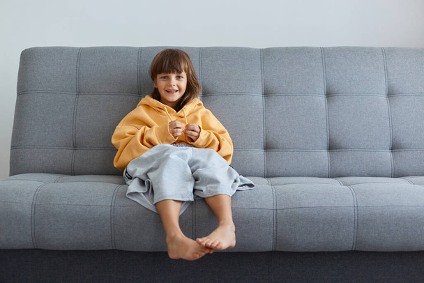 Portrait of dark haired little girl wearing oversized yellow hoodie and gray short sitting on cough and looking at camera, posing indoor alone, expressing positive emotions. - Fotoğraf, Görsel