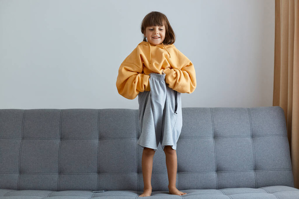 Full lengh portrait of Caucasian little girl wearing oversized yellow hoodie and gray short standing on sofa, holding big clothing and looking at camera with funny facial expression. - Foto, immagini
