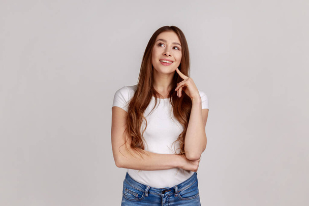 Portrait of adorable dreamy woman standing looking away, keeping finger on cheek, dreaming, making wish, love, wearing white T-shirt. Indoor studio shot isolated on gray background. - Photo, image
