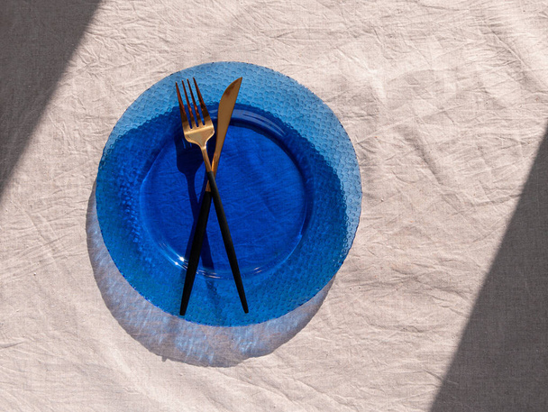 Tischdekoration leerer blauer Glasteller mit Gabelmesser auf Leinentücherdeckel bei Tageslicht und harten Schatten. Festlicher Ort Natürliche Baumwollkerne stilvolles Geschirr minimale Wohnkultur Landschaft Ästhetik - Foto, Bild
