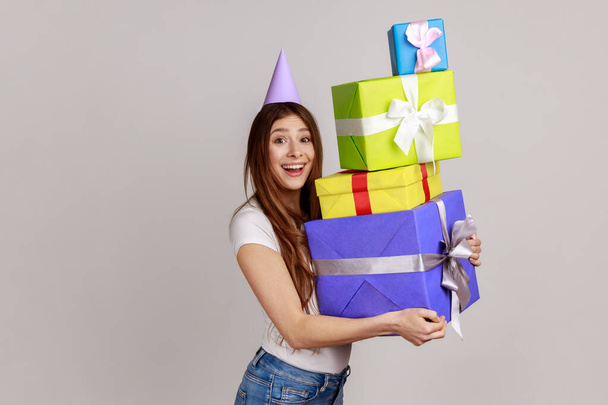 Portrait of joyful young woman wearing party cone, holding mount of gift boxes, enjoying many best presents, celebrating birthday, wearing white T-shirt. Indoor studio shot isolated on gray background - Foto, Imagen
