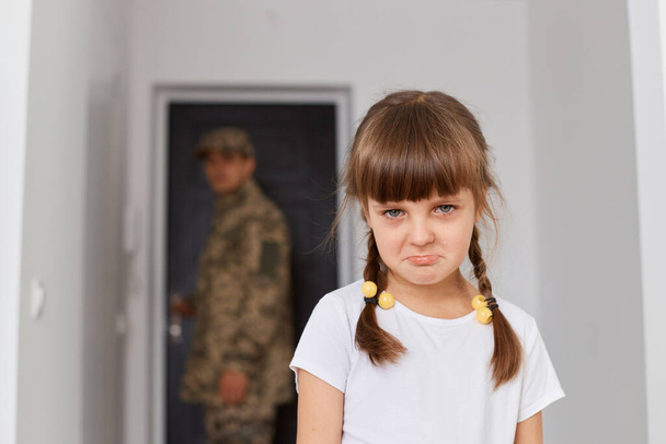 Retrato de una niña infeliz vistiendo una camiseta blanca con labios de puchero de pie, mirando a la cámara, molestando a su padre militar yendo a la guerra o al ejército, un hombre borroso de pie en la puerta. - Foto, imagen
