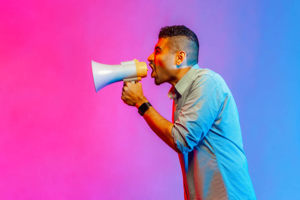 Side view portrait of angry man in shirt loudly speaking screaming holding megaphone, announcing important message. Indoor studio shot isolated on colorful neon light background. - Photo, image