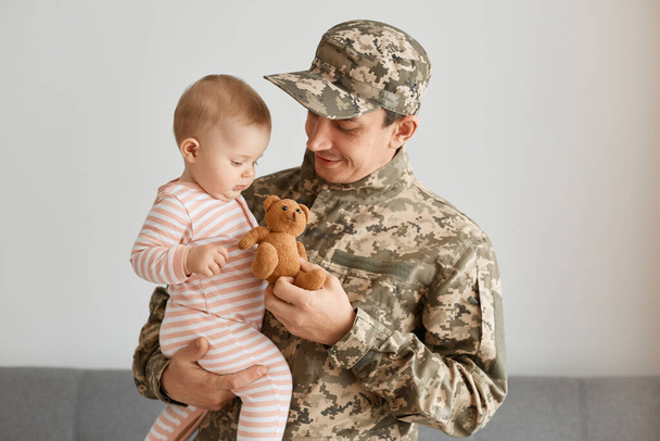 Portrait of soldier man wearing camouflage holding his infant daughter and giving her little teddy bear toy, present after returning home from army or war. - Photo, Image