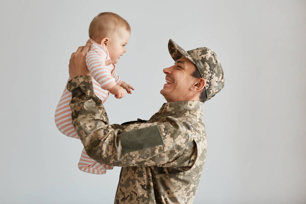 Side view portrait of young adult soldier man in camouflage uniform and cap throwing his toddler daughter on white background indoor , male returning home from war, being happy to see his kid. - Photo, image