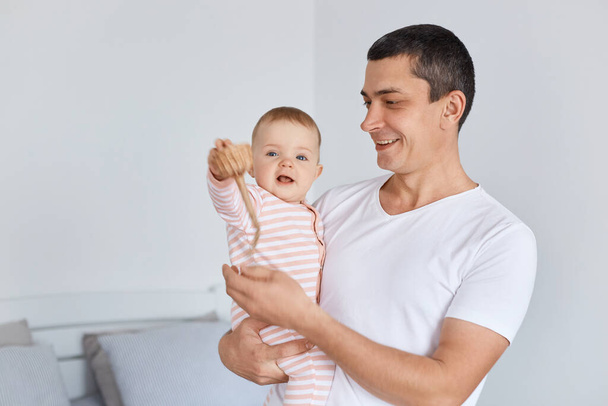 Smiling positive man holding his newborn baby daughter, man expressing happiness, spending time with hid child, posing indoor at home, cute kid raised arm and looking at camera. - Photo, Image