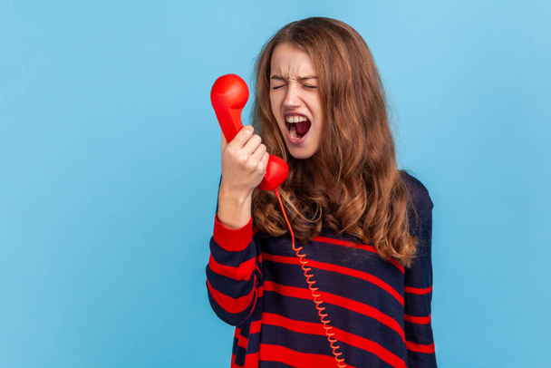 Angry woman wearing striped casual style sweater, screaming while talking on landline phone, conversation with annoying client on call center. Indoor studio shot isolated on blue background. - Photo, Image