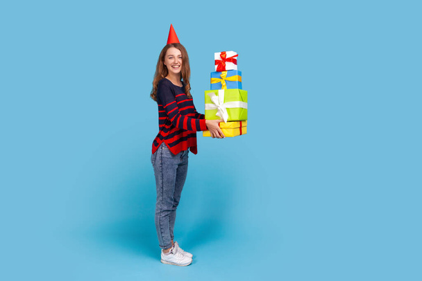 Full length profile portrait of woman wearing sweater and party cone holding many boxes with birthday presents, looking at camera with pleasant smile. Indoor studio shot isolated on blue background - Fotografie, Obrázek