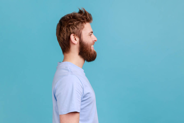 Side view of bearded man looking at camera with toothy smile and happy facial expression, being in good mood, rejoicing great results. Indoor studio shot isolated on blue background. - Foto, Imagem
