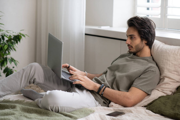 Молодий бізнесмен в повсякденному одязі лежить на ліжку і друкує на клавіатурі ноутбука
 - Фото, зображення