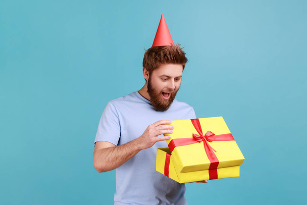 Man with party cone hat holding opened gift box and looking at present with excited look, unpacking present, enjoying awesome birthday surprise. Indoor studio shot isolated on blue background. - Photo, image