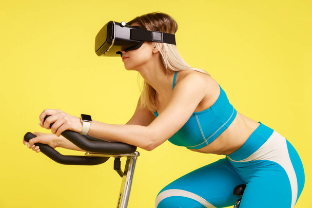 Side view portrait of fit grinning woman in blue sportswear exercising on sports machine wearing VR-headset, having cardio workout. Indoor studio shot isolated on yellow background. - Fotó, kép