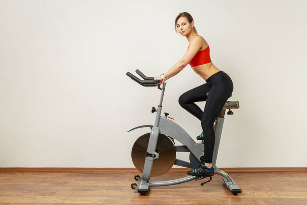 Side view portrait of slim athletic woman with perfect body shape training on bicycle, using sport equipment for workout, wearing sports tights and top. Indoor studio shot on gray wall background. - Zdjęcie, obraz