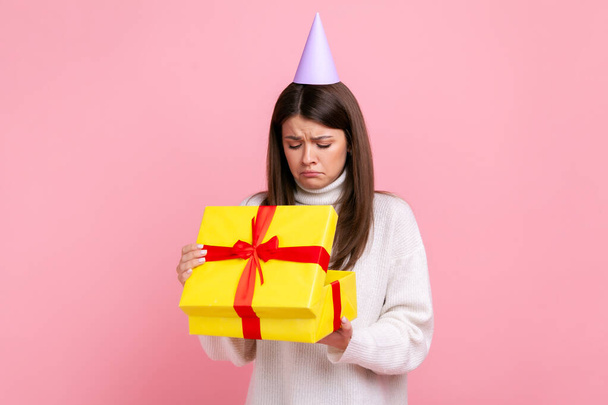 Disappointed female in party cone looking inside wrapped present box, open gift, unpleasant surprise, wearing white casual style sweater. Indoor studio shot isolated on pink background. - Photo, image