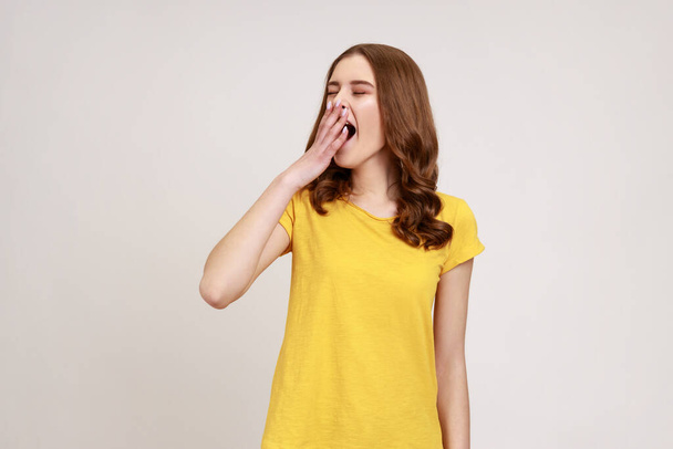 Portrait of sleepy teenager girl with wavy hair in casual yellow t-shirt yawning with closed eyes and covering mouth, feeling exhausted need rest. Indoor studio shot isolated on gray background. - Фото, зображення